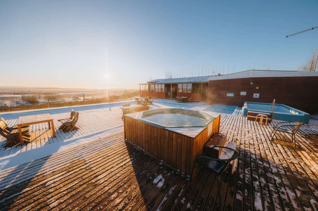 Sunlit deck with snow-covered wooden planks, hot tub, and lounge chairs in foreground. A modern building and vast landscape under a clear blue sky.