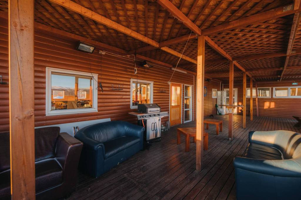 Wooden deck with sofas, tables, and a grill under a warm-toned wooden roof. Windows and doors line the cabin-style walls.