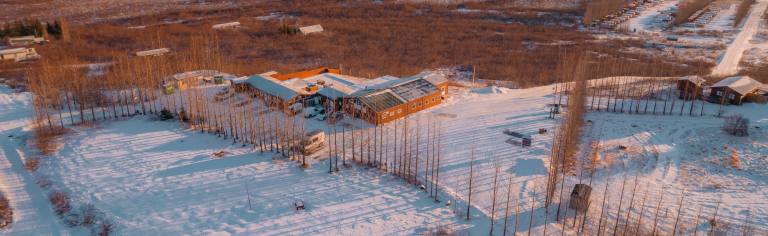 Aerial view of a snow-covered landscape with scattered trees and a cluster of rustic buildings. The warm sunset casts long shadows.
