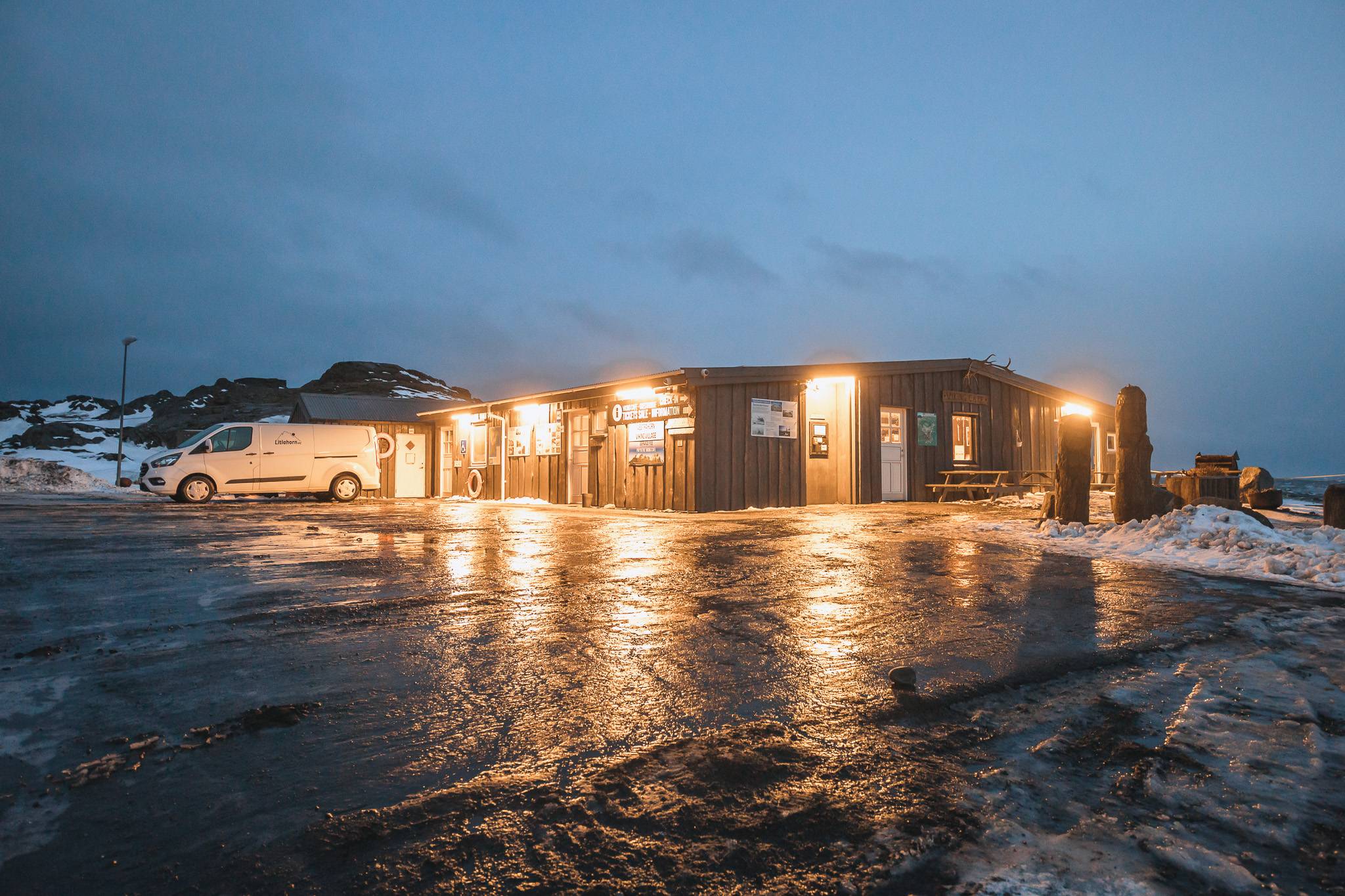 A warmly lit cabin sits in a snowy, icy landscape at dusk, reflecting light on the wet ground. A van is parked nearby under a cloudy evening sky.