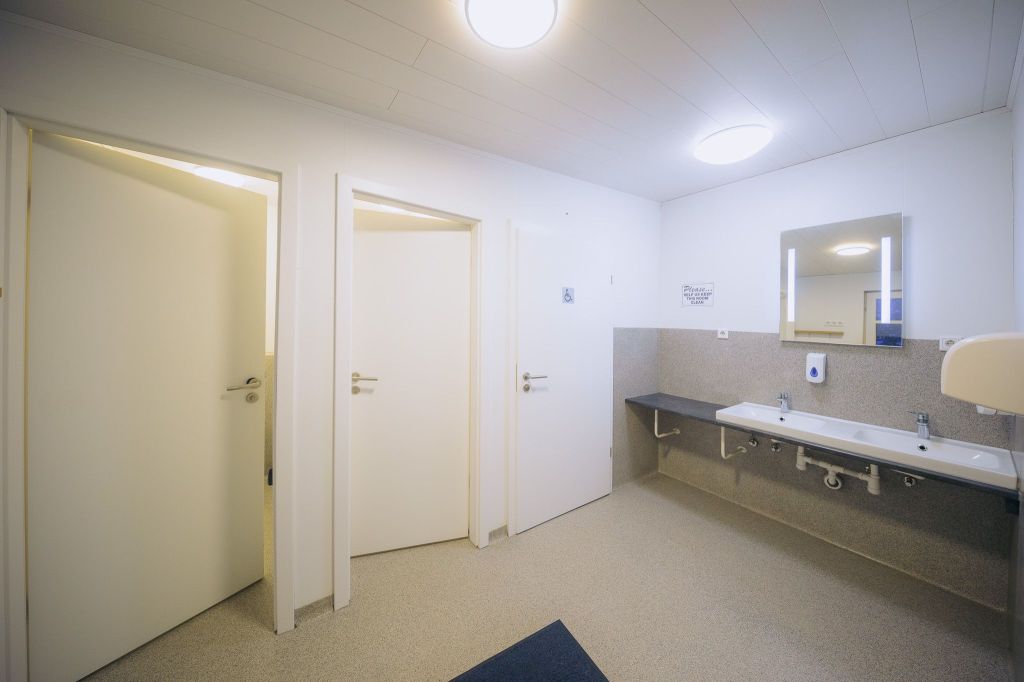 A clean, bright public restroom with three closed stalls on the left. On the right, a long counter with two sinks, a mirror, and a soap dispenser.