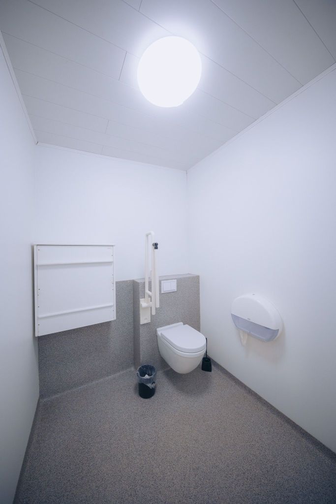 Minimalist, clean restroom with white walls and a gray floor. Features a wall-mounted toilet, support rail, waste bin, and paper dispenser under bright lighting.