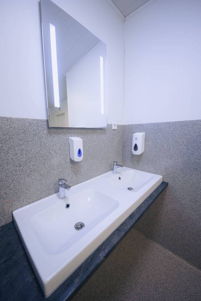 A clean, modern bathroom features a double sink with chrome faucets, a large mirror, and two wall-mounted soap dispensers on a light grey wall.