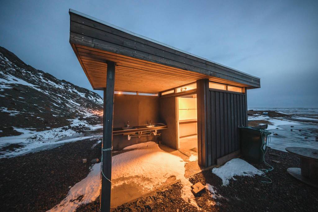 A small, modern cabin with an open section and warm interior light stands on a snowy landscape at dusk.