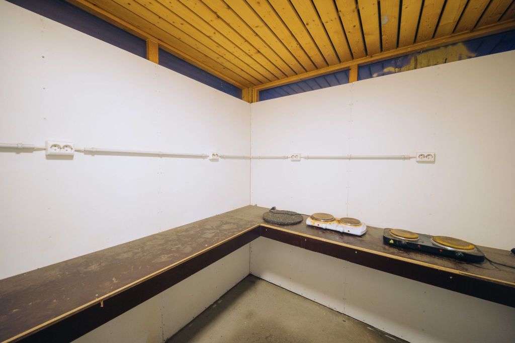 Interior of an empty, wood-ceilinged room with white walls and a corner countertop. Two electric hot plates rest on the counter.