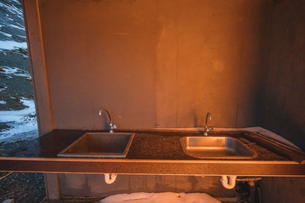 Outdoor sink area with two metal basins and faucets on a dark countertop, illuminated by warm light. Snow is visible on the ground and nearby surfaces.