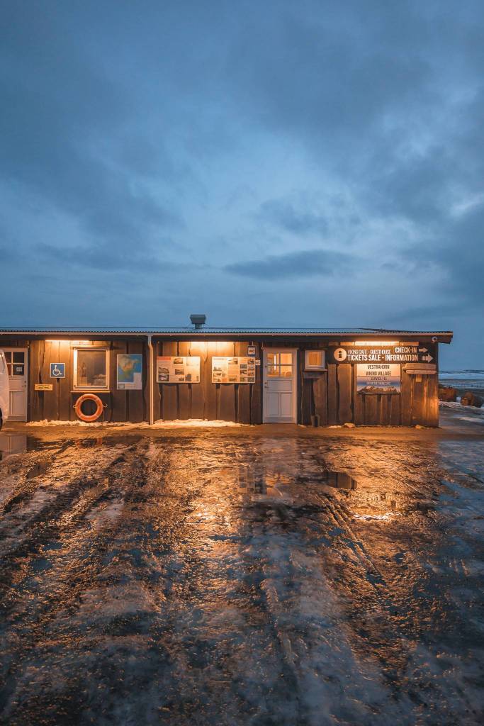 A dimly lit wooden building with ticket and information signs stands against a dusky sky. Wet ground reflects warm lights.