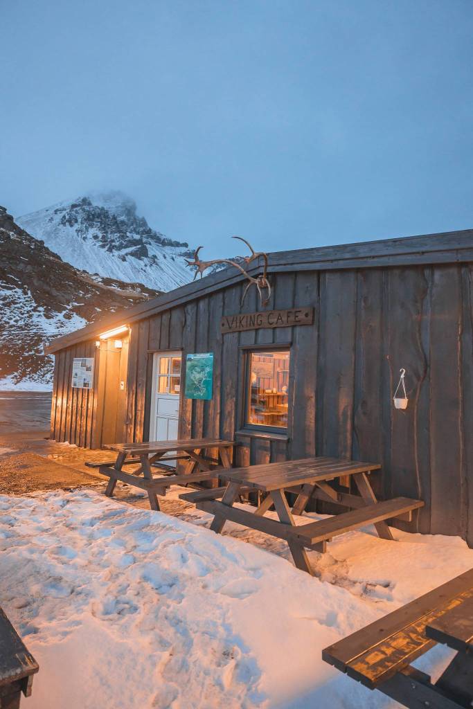 Wooden cabin labeled "Viking Café" with warm lights, snow-covered picnic tables, and a mountain backdrop.