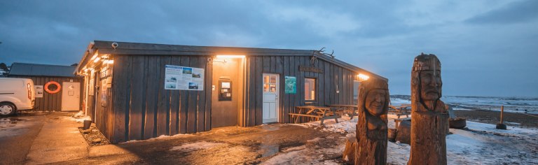 A cozy seaside building with warm lights at dusk. Wooden sculptures stand outside on a snowy path.