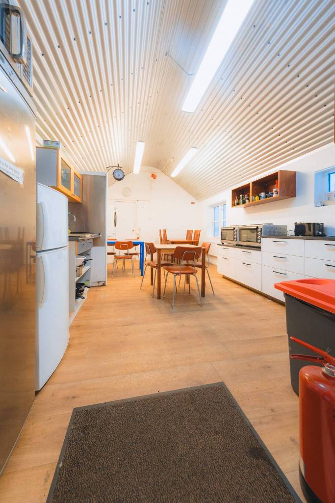 Wood-floored kitchen with a high, corrugated ceiling. Features white cabinets, wooden table with chairs, fridge, and fire extinguisher.