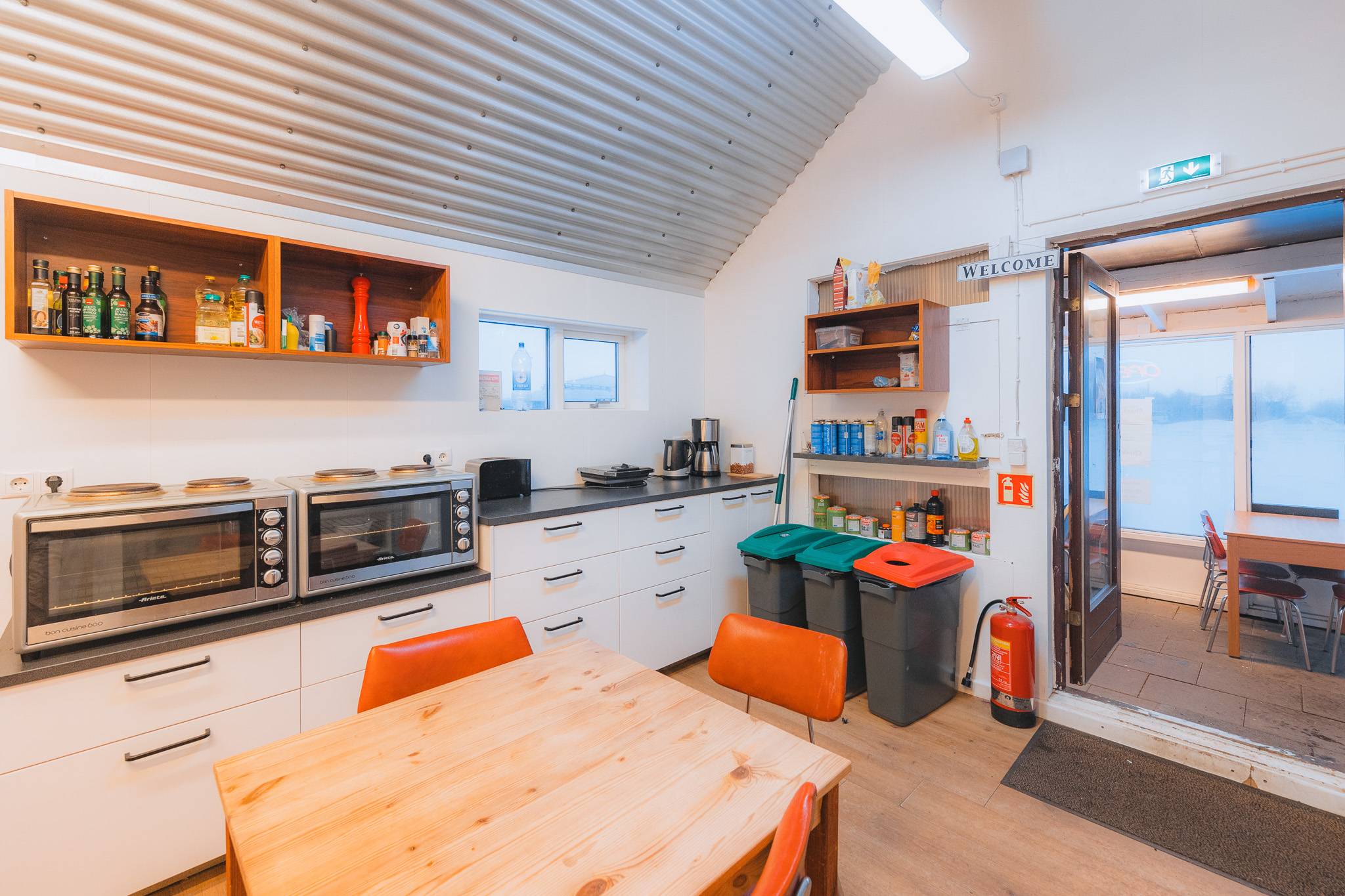 Cozy kitchen with wooden table and orange chairs, double ovens, shelves with bottles and cans. Recycling bins and fire extinguisher near the door.