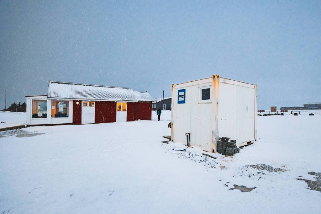 Snowy landscape with a white portable restroom in the foreground. A red building with lit windows is in the background, under a gray, overcast sky.