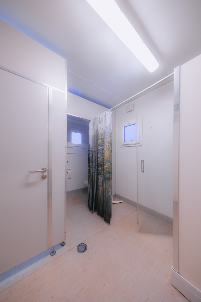 Simple bathroom with white walls and floor, a fluorescent light overhead, a shower area with a forest-patterned curtain, and a small window.