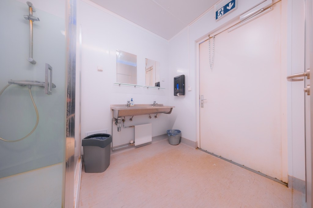 A clean public bathroom with a shower on the left, a long sink with a mirror and soap dispenser in the center, and an exit door on the right.