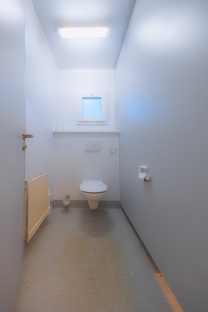 Narrow, minimalist bathroom with white walls and ceiling. A toilet is centered under a small window. A radiator and toilet paper holder flank it.