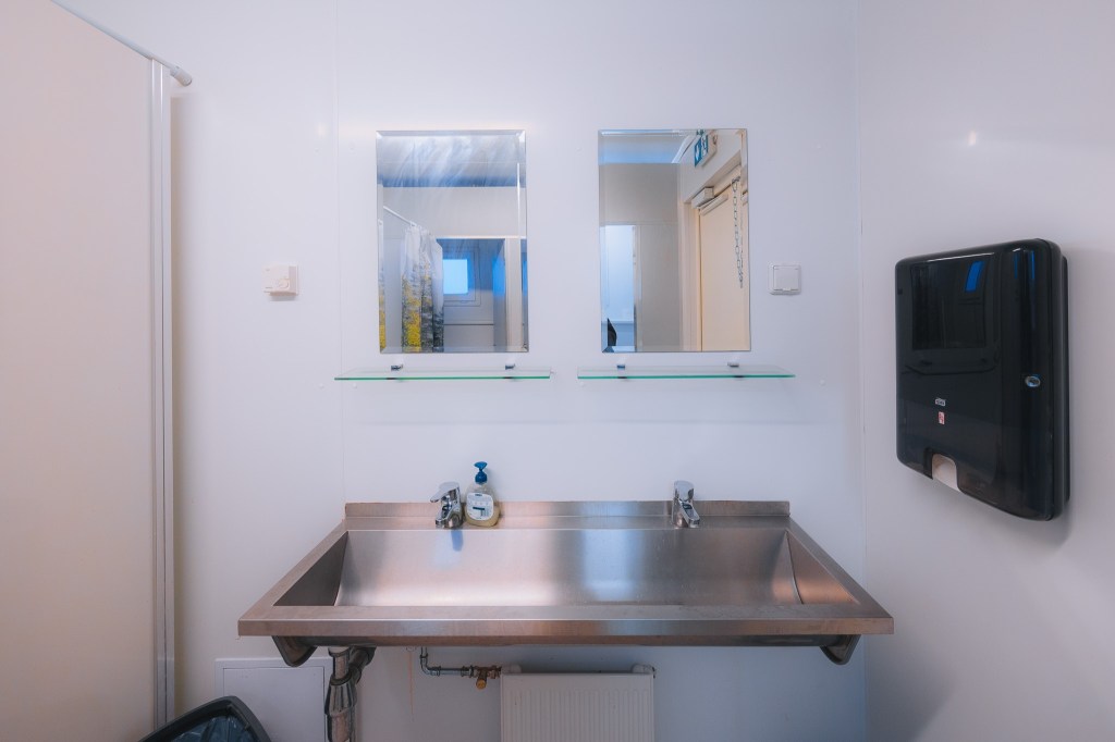 Industrial-style bathroom with a stainless steel double sink, two mirrors, soap dispenser, and paper towel holder.