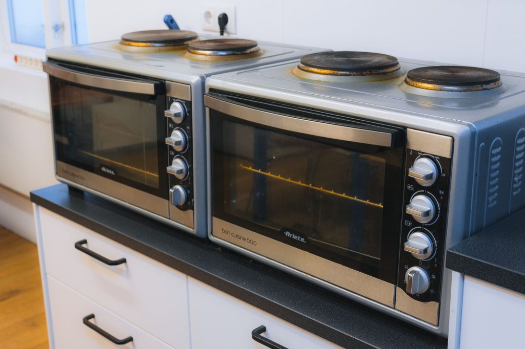 Two stainless steel countertop ovens with stove tops are placed side by side on a dark counter in a bright kitchen.
