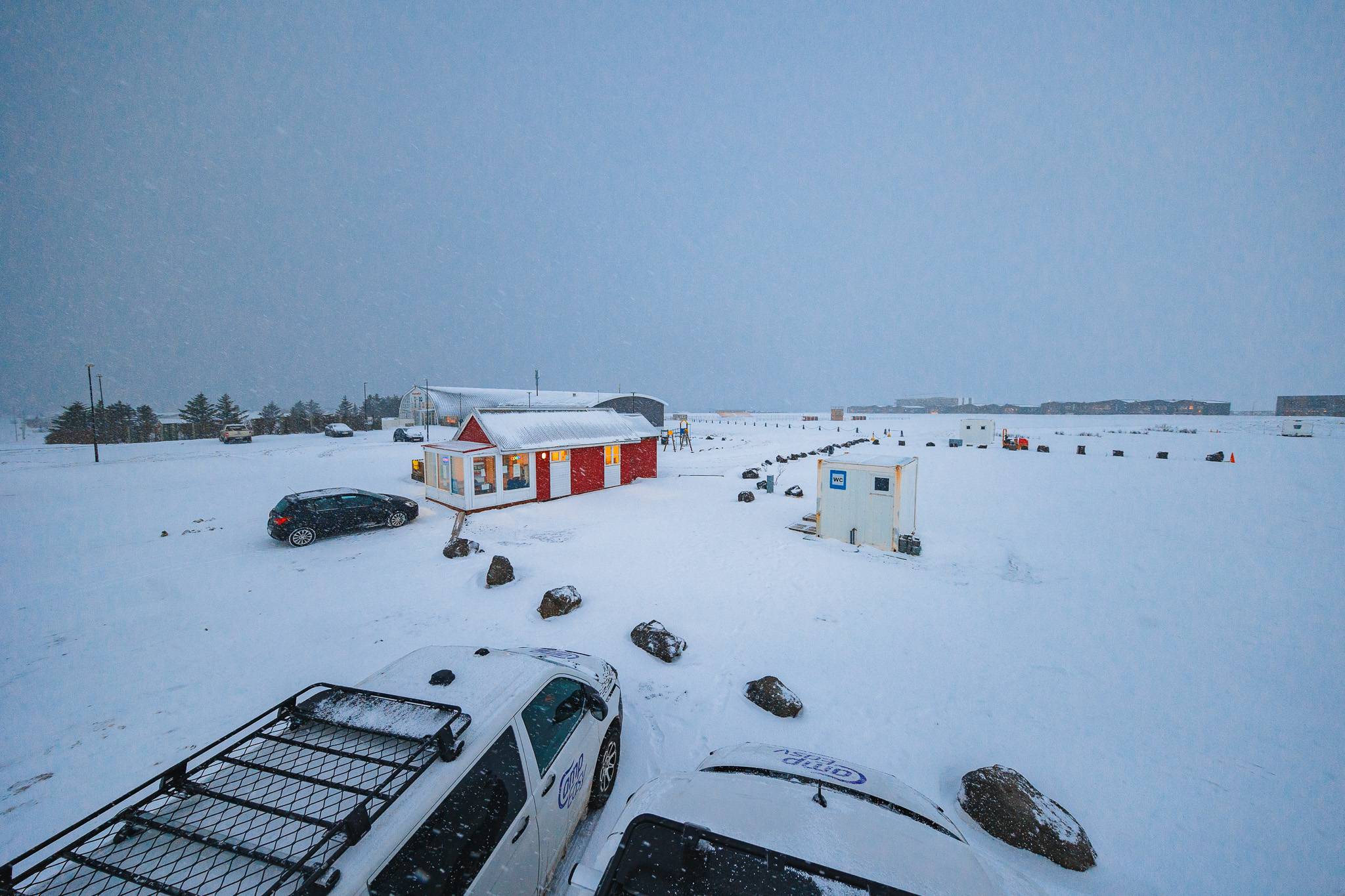 A snowy landscape with a bright red and white building in the center, surrounded by snow-covered ground, cars, rocks, and a gray overcast sky.