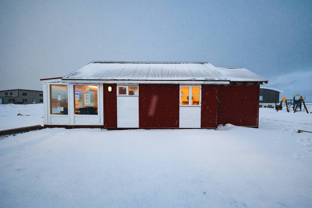 A cozy red building with white doors and glowing windows sits on a snowy landscape at dusk. A swing set is visible in the background.