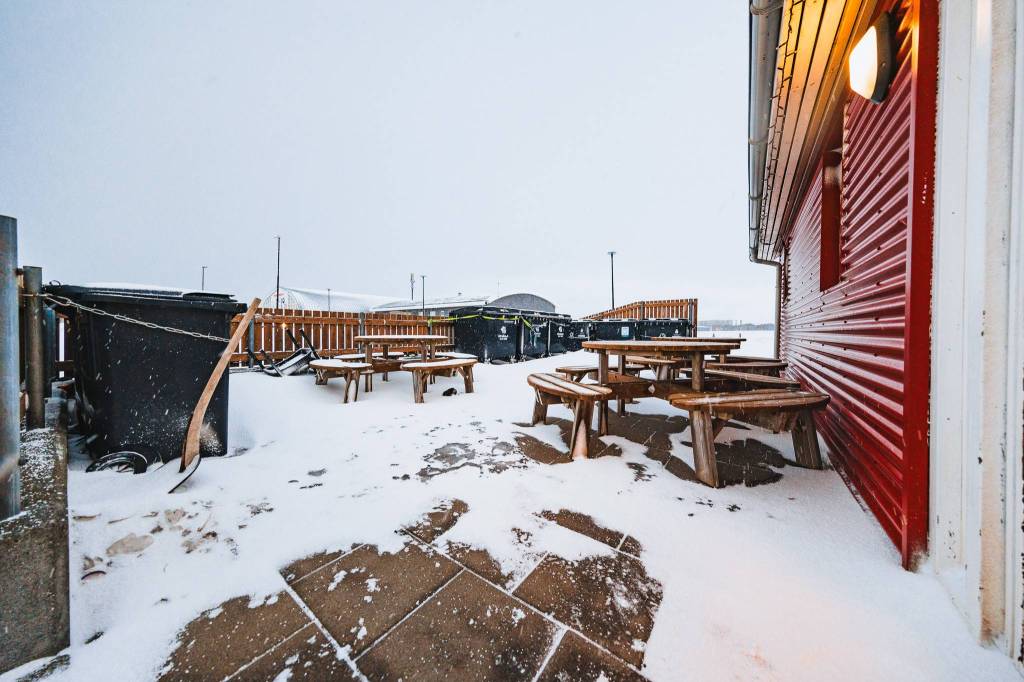 Snow-covered outdoor seating area with wooden picnic tables beside a building with a red corrugated exterior.