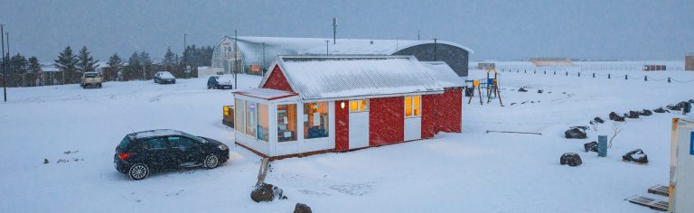 Cozy red cabin with glowing windows in snowy landscape, parked car nearby, overcast sky, and scattered buildings in the background.