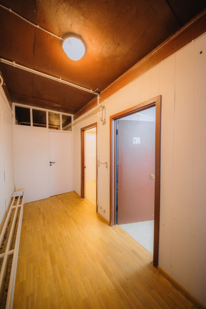 A narrow hallway with wooden flooring and white walls is lit by a ceiling lamp. Two open doors on the right lead to adjacent rooms, one marked with a restroom sign.