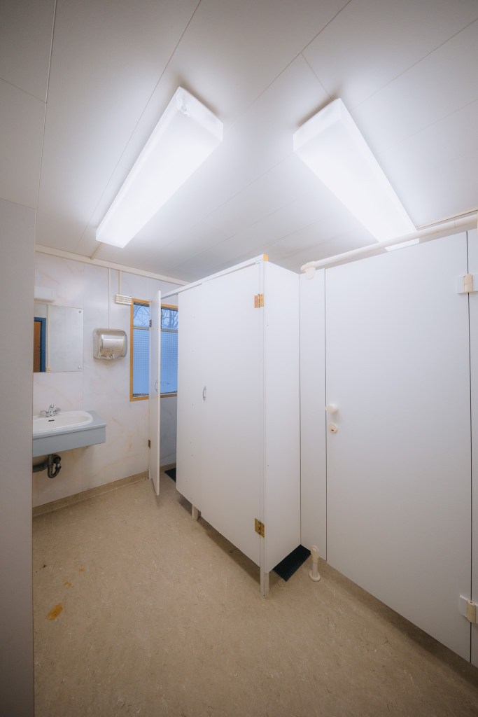 Public restroom interior with white stalls, beige flooring, and fluorescent ceiling lights. A sink with a mirror is on the left.