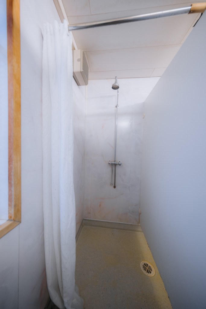 Narrow shower stall with white curtain, adjustable showerhead, and simple gray walls.