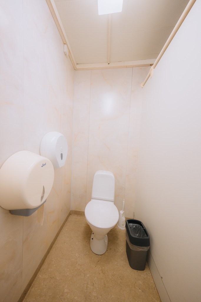 A narrow shower space with white walls, showerhead, and a white curtain. Shelves are mounted on the wall, and a small wooden stool is in the foreground.