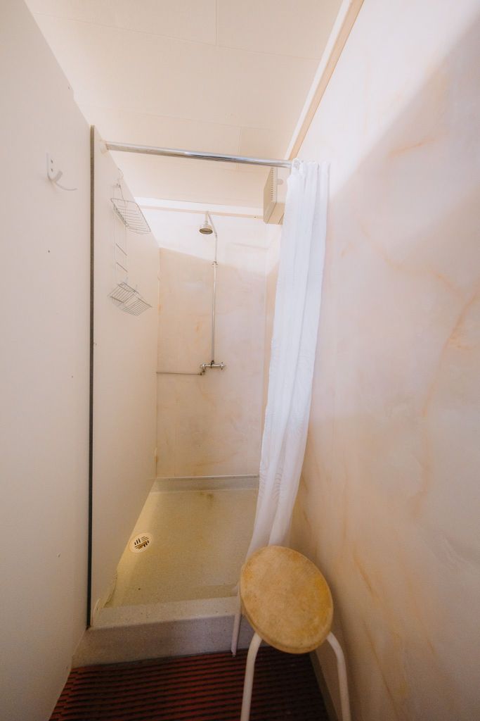 A narrow shower space with white walls, showerhead, and a white curtain. Shelves are mounted on the wall, and a small wooden stool is in the foreground.