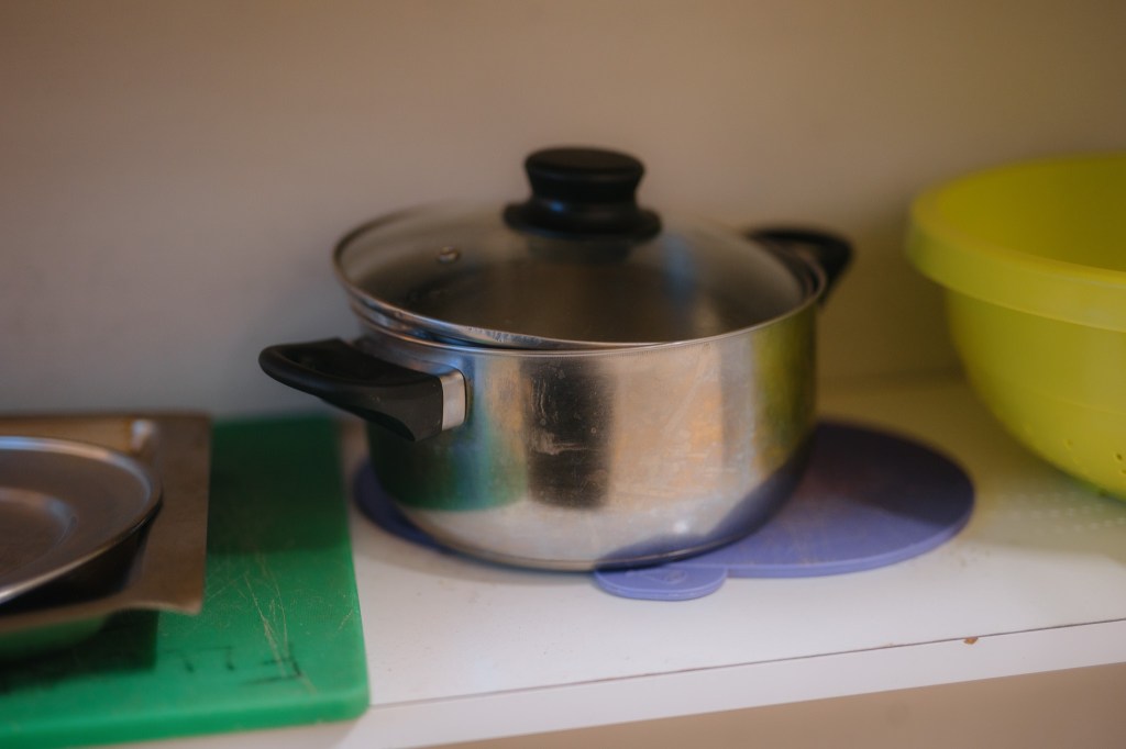 A stainless steel pot with a glass lid sits on a purple mat in a kitchen cupboard, alongside a green cutting board and a yellow bowl.
