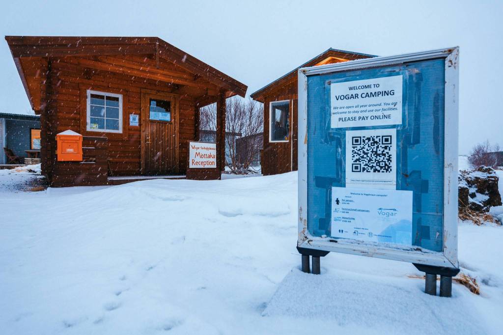 Cozy wooden cabins in a snowy landscape, with a sign for Vogar Camping's reception. Another sign displays a QR code for online payment.