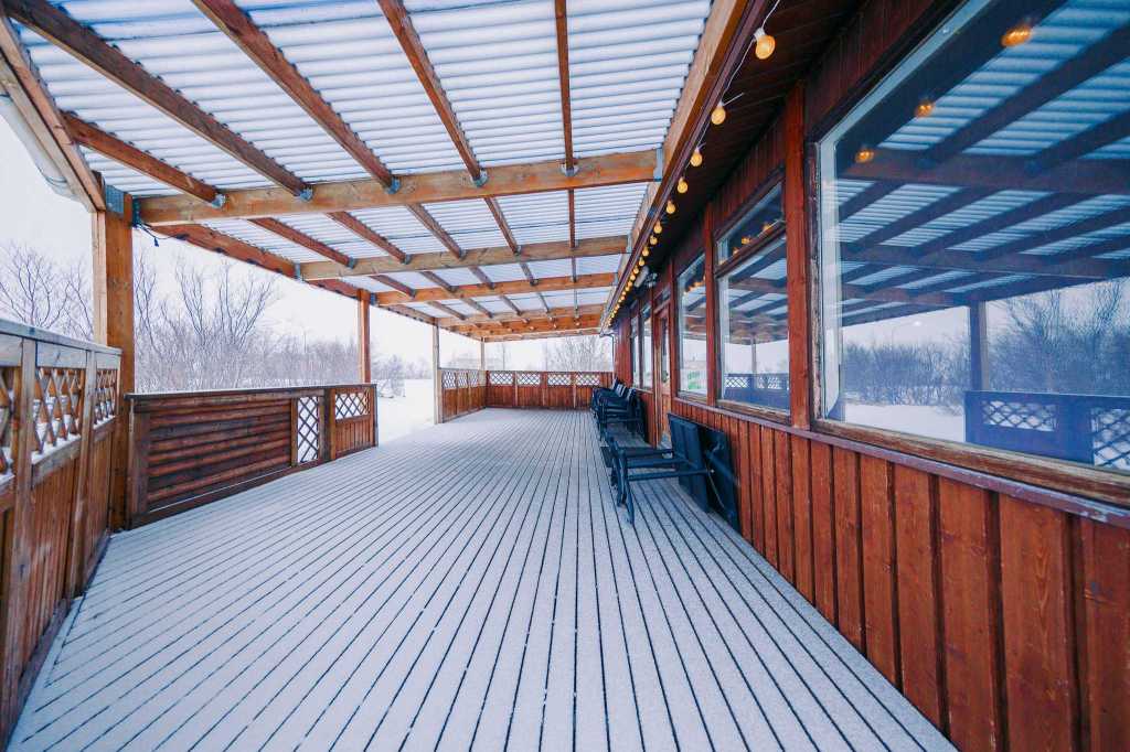 Wooden deck covered in snow, with string lights under a pergola roof. Several black chairs line the right side, overlooking a snowy landscape.