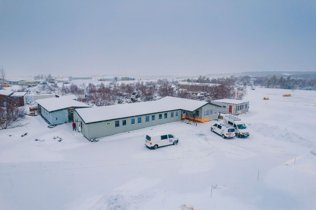 A snowy landscape features a long, gray building surrounded by trees. Three white campervans are parked outside