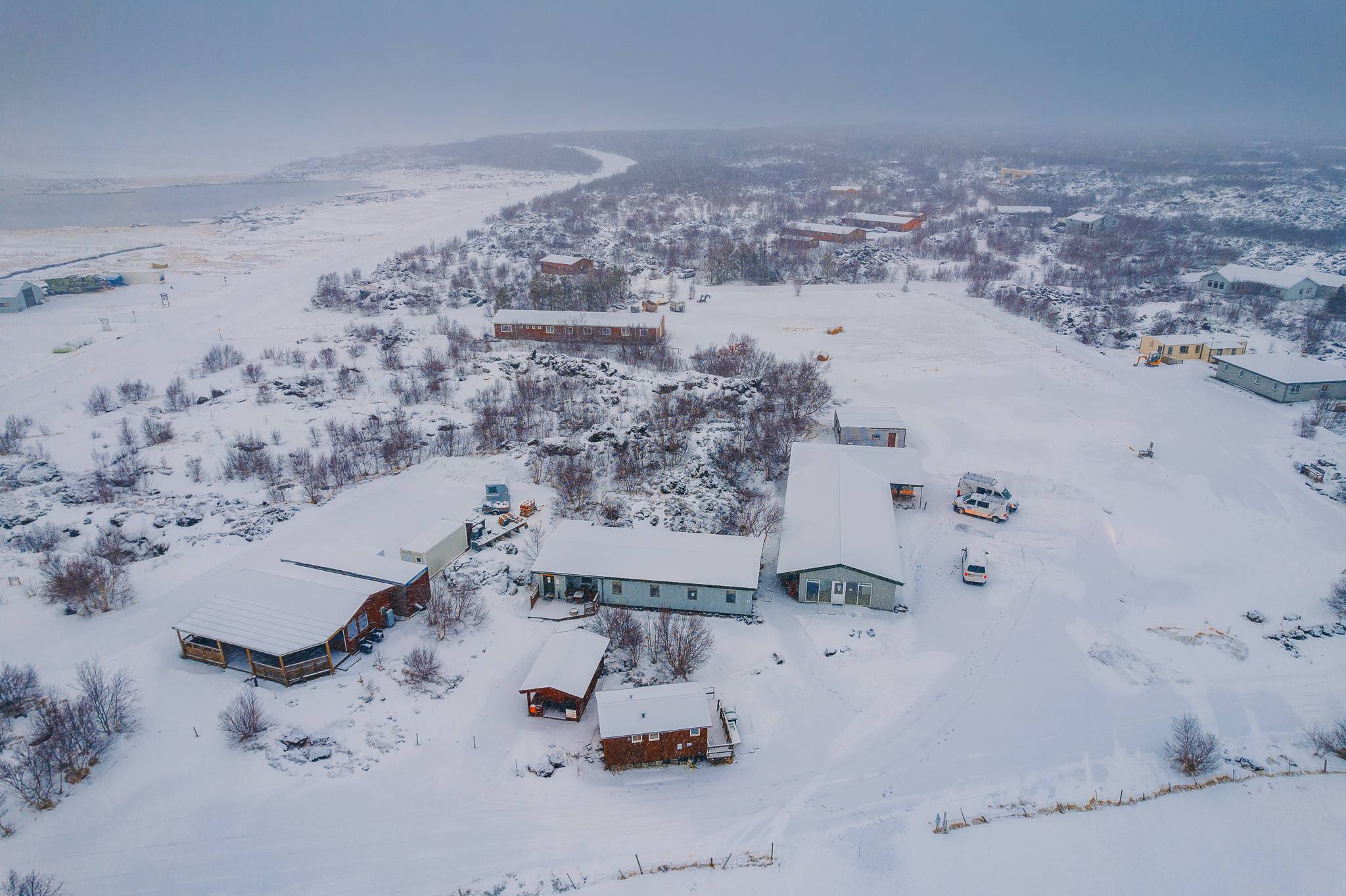 Aerial view of a snowy landscape with scattered buildings and trees. Overcast sky and distant waterway.