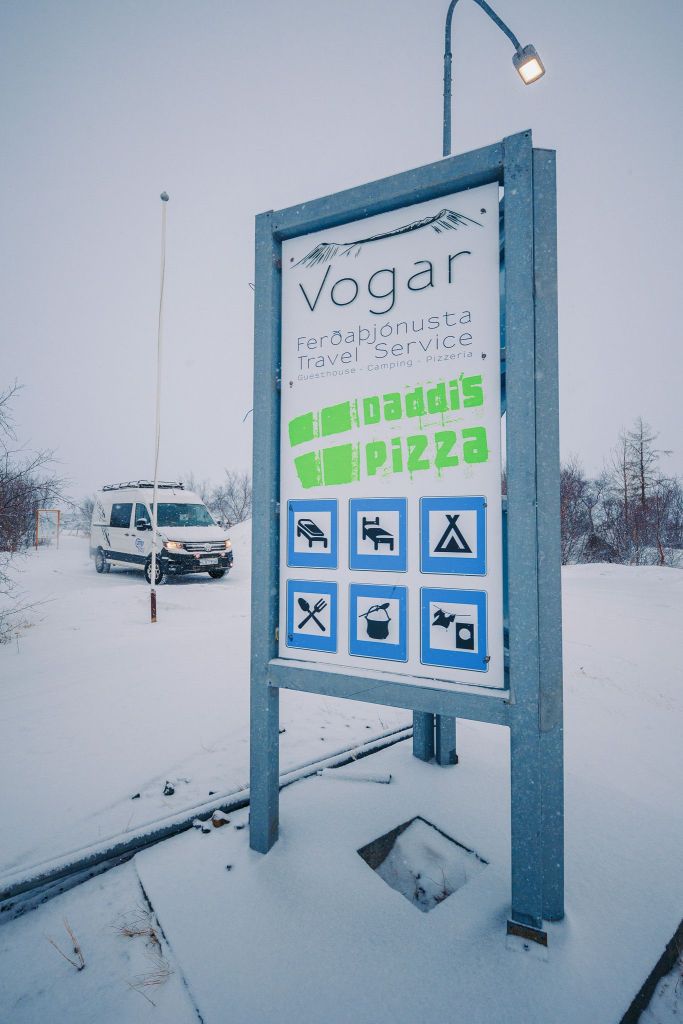 Roadside sign in snowy landscape reads "Vogar Travel Service, Daddi's Pizza" with symbols for amenities. A parked campervan is visible in the background.