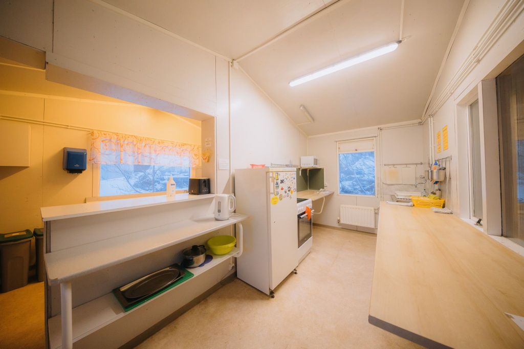 Simple kitchen with white walls and beige floor, featuring a fridge with magnets, shelves with utensils, and a window with a snowy view and soft lighting.