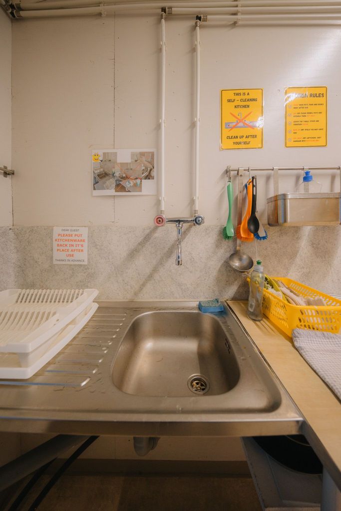 A communal kitchen sink with utensils on wall hooks. Signs urge cleaning up. Nearby are a dish rack, yellow basket, and cleaning supplies.
