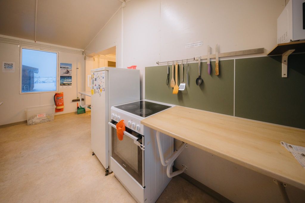 Simple kitchen with a stove, refrigerator, and hanging utensils. A window shows a snowy scene.