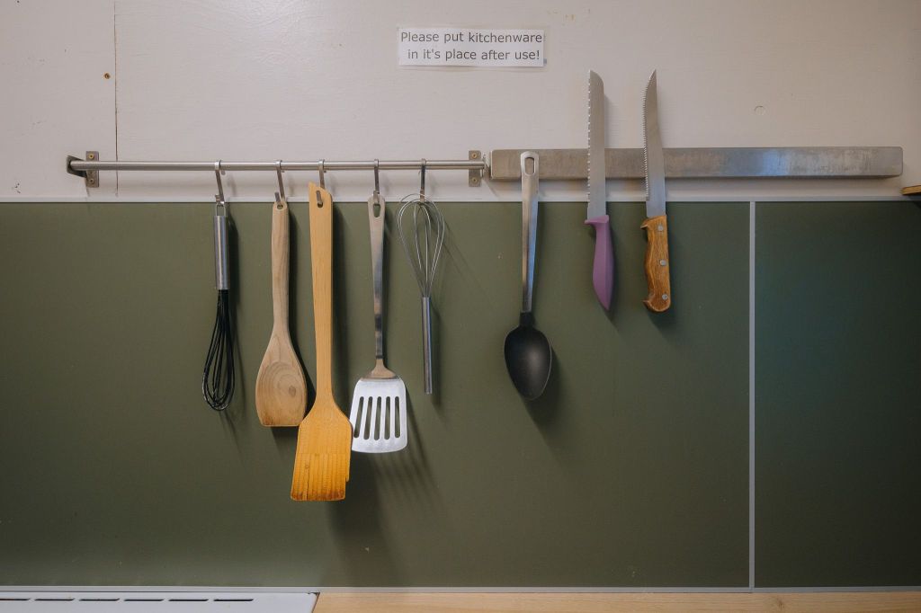 A row of kitchen utensils hangs neatly on a wall-mounted rack against a green backsplash. Above is a sign: "Please put kitchenware in its place after use!"