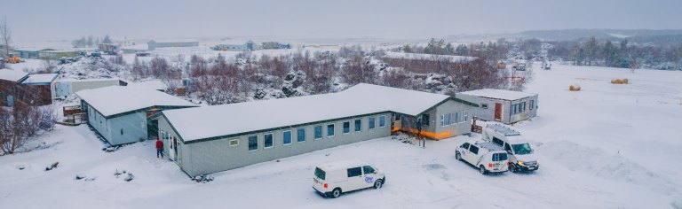 A snowy landscape features a long, gray building surrounded by trees. Three white campervans are parked outside